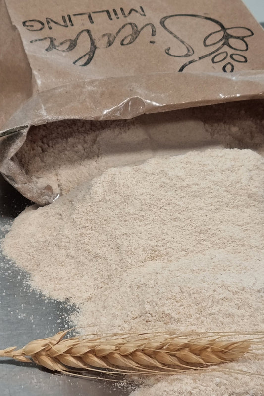 A close-up of a brown paper bag labeled 'Sieda Milling' next to a pile of light beige flour and a single wheat ear on a dark surface.