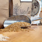 A close-up of a milling process showing a metal scoop filled with light beige flour in the foreground, scattered whole grains in the center, and brown paper bags labeled 'Sieda Milling' in the background on a wooden table.