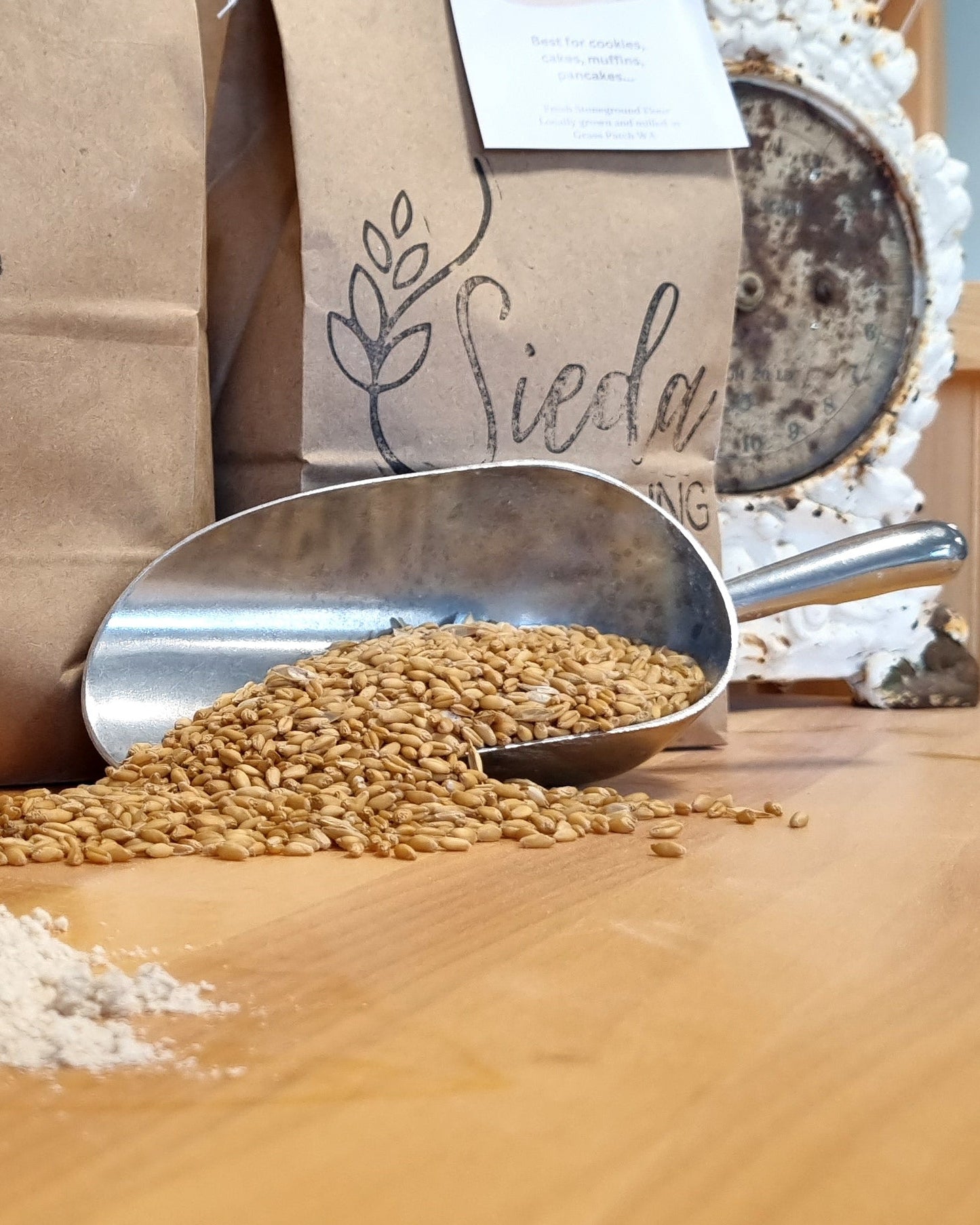 A close-up of a milling process showing a metal scoop filled with light beige flour in the foreground, scattered whole grains in the center, and brown paper bags labeled 'Sieda Milling' in the background on a wooden table.