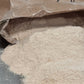 A close-up of a brown paper bag labeled 'Sieda Milling' next to a pile of light beige flour and a single wheat ear on a dark surface.