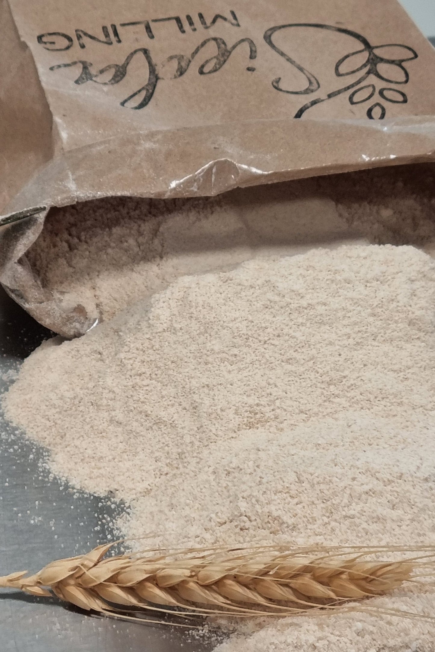 A close-up of a brown paper bag labeled 'Sieda Milling' next to a pile of light beige flour and a single wheat ear on a dark surface.
