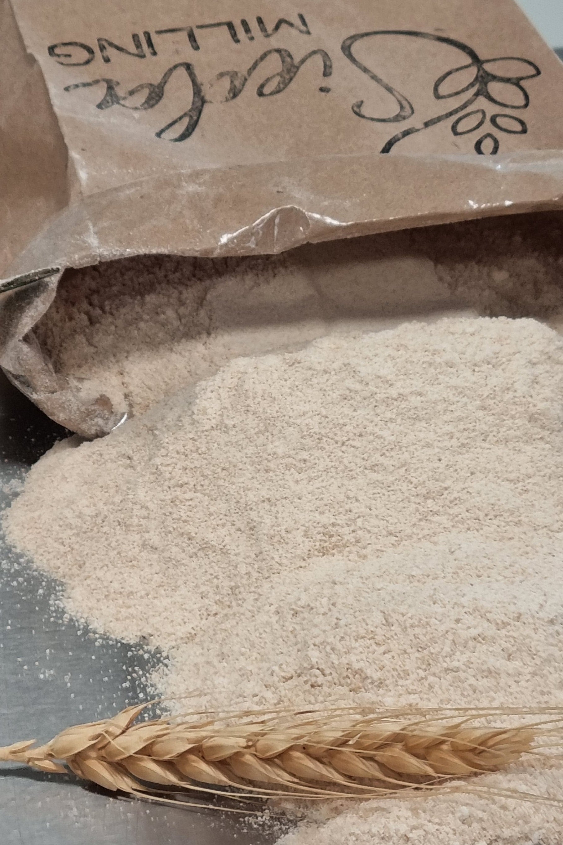 A close-up of a brown paper bag labeled 'Sieda Milling' next to a pile of light beige flour and a single wheat ear on a dark surface.