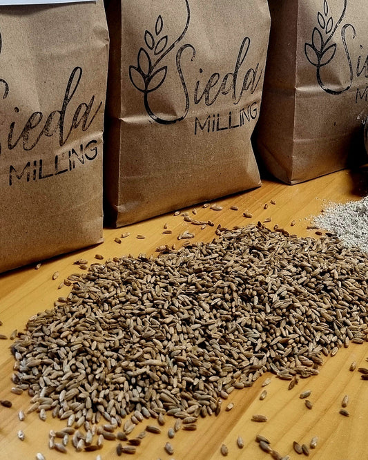 Brown paper bags labeled 'Sieda Milling' are arranged on a wooden table with loose rye grains and flour spilled in front.
